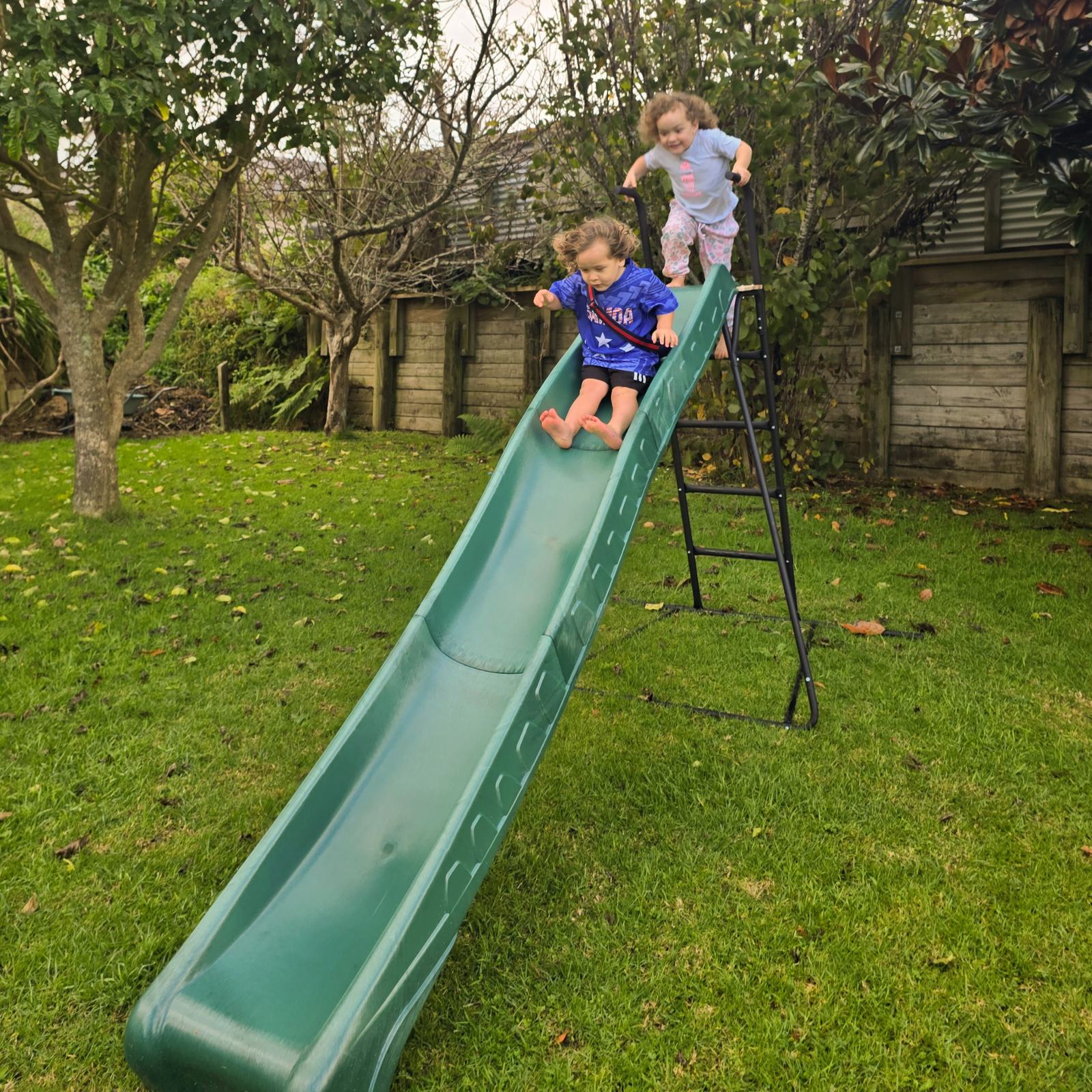 2 children having great fun sliding down a green slide in NZ backyard