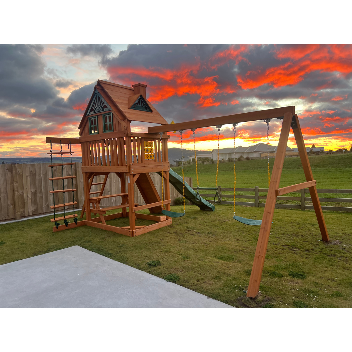 A wooden outdoor playground with a house tower, ladder, slide, and swing set, shown on a grassy backyard under a dramatic orange and gray sunset sky.