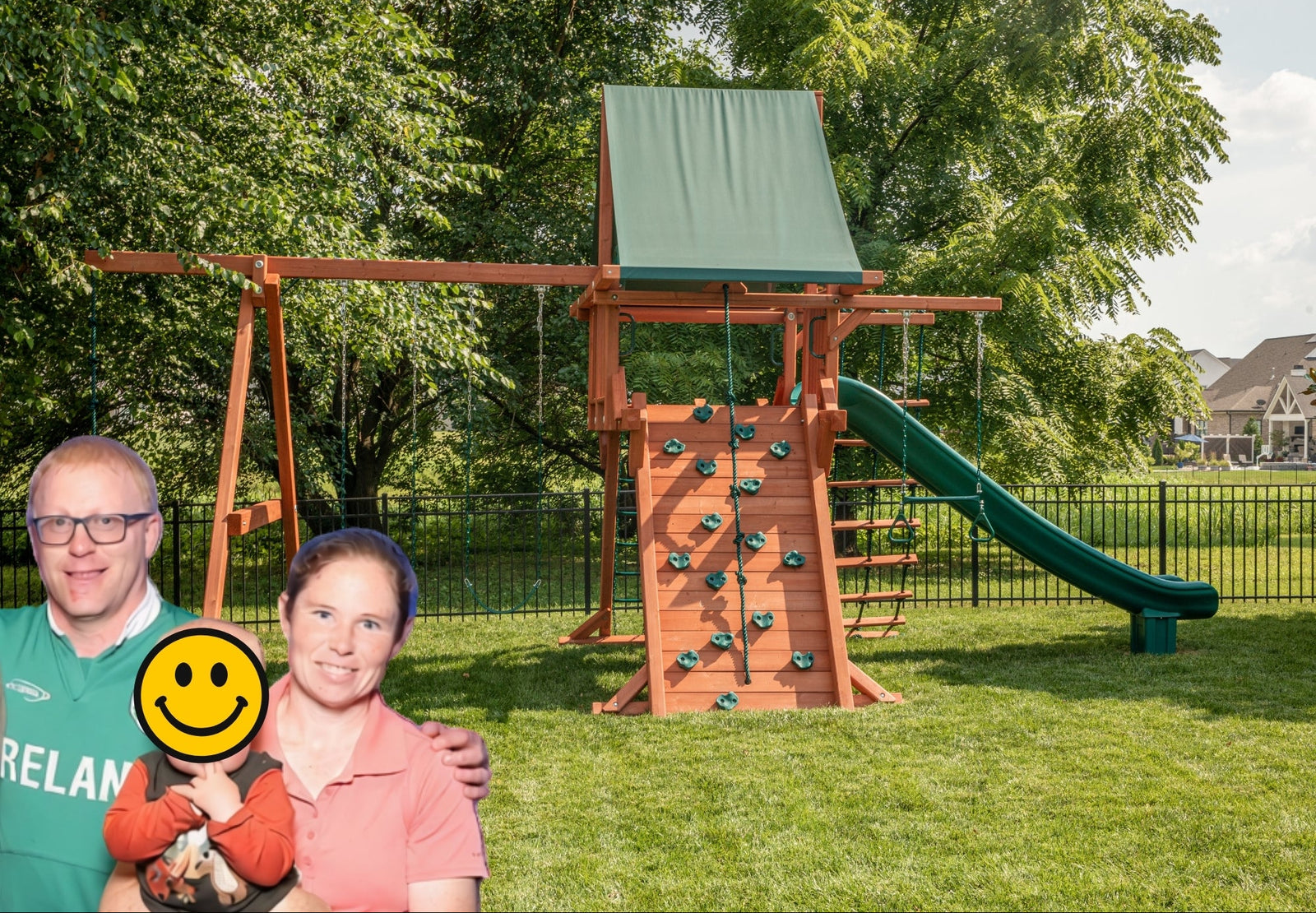 A large wooden backyard playset with a green slide, climbing wall, and swing set, shown on a grassy lawn with trees in the background. Kiwiplay owners are pictured with their child in front of the playground