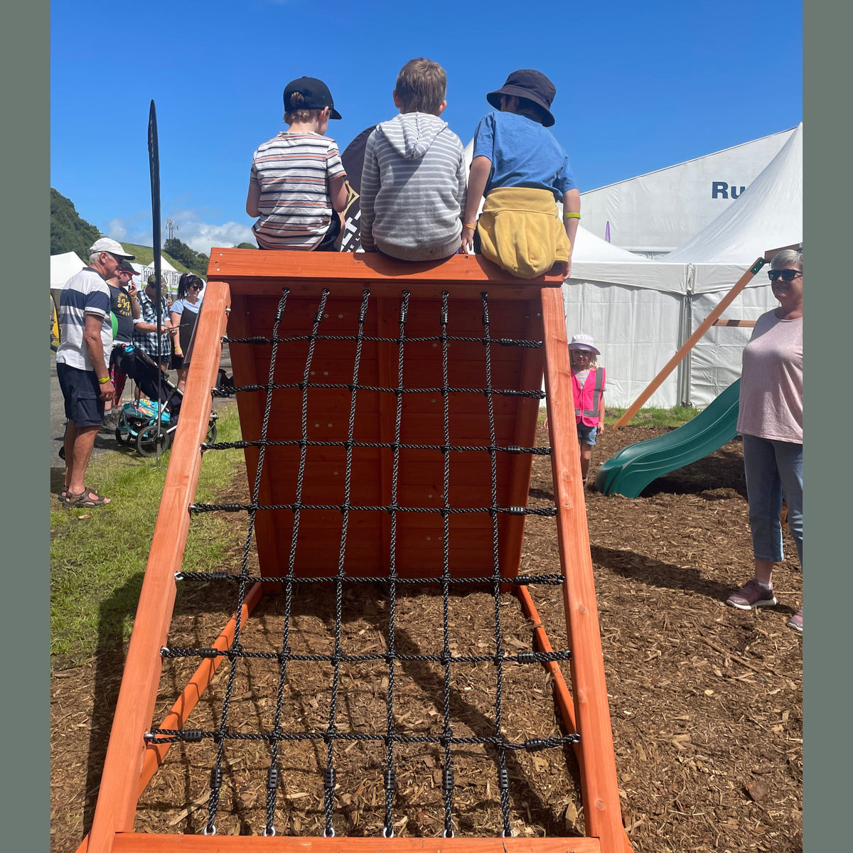 A wooden outdoor climbing structure with a rope net front and three children sitting on top.