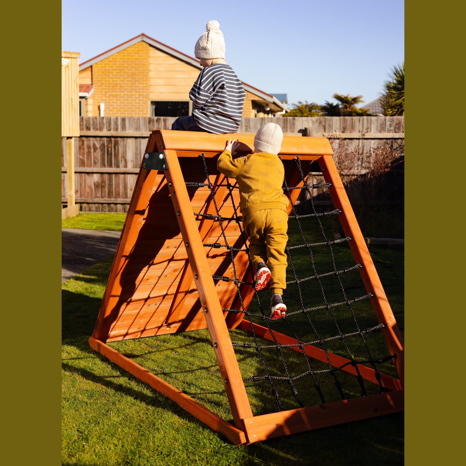A children's wooden A-frame climbing wall with black rope netting, shown outdoors on a grassy backyard. One child is climbing the wall while another sits on top.
