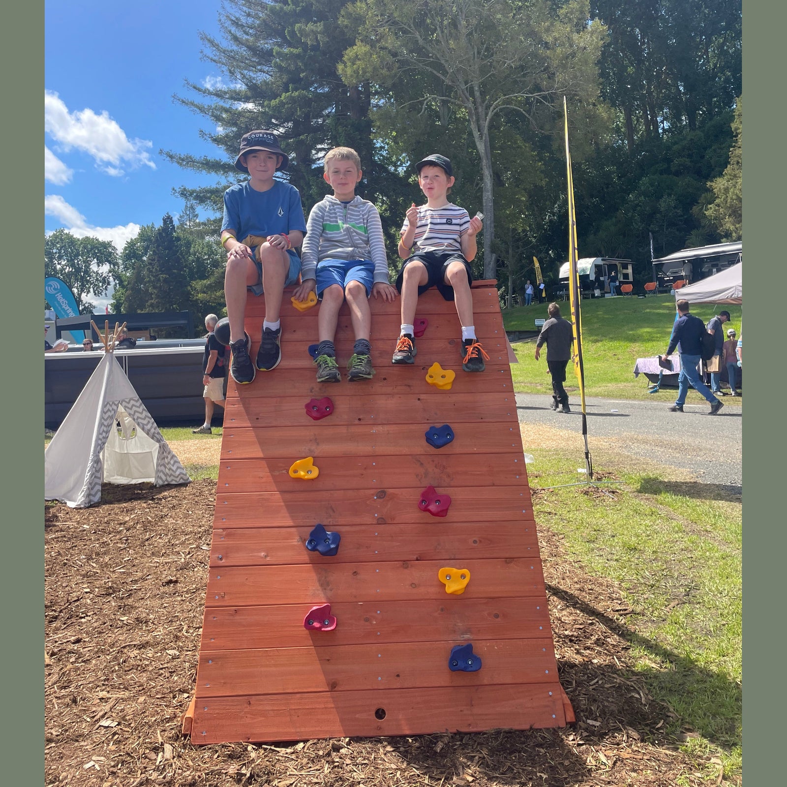 Three children sitting on top of a wooden climbing wall with colorful handholds in an outdoor park setting with trees and a blue sky.