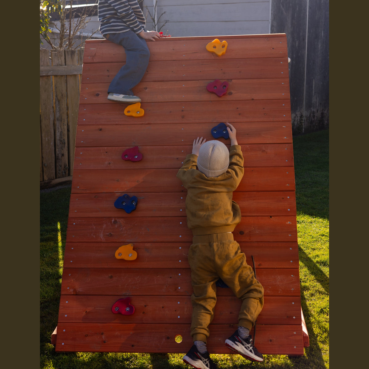 Two children playing on a wooden climbing wall outdoors in a backyard. One child is sitting at the top of the wall, and the other is climbing up the wall using colorful hand and foot holds.