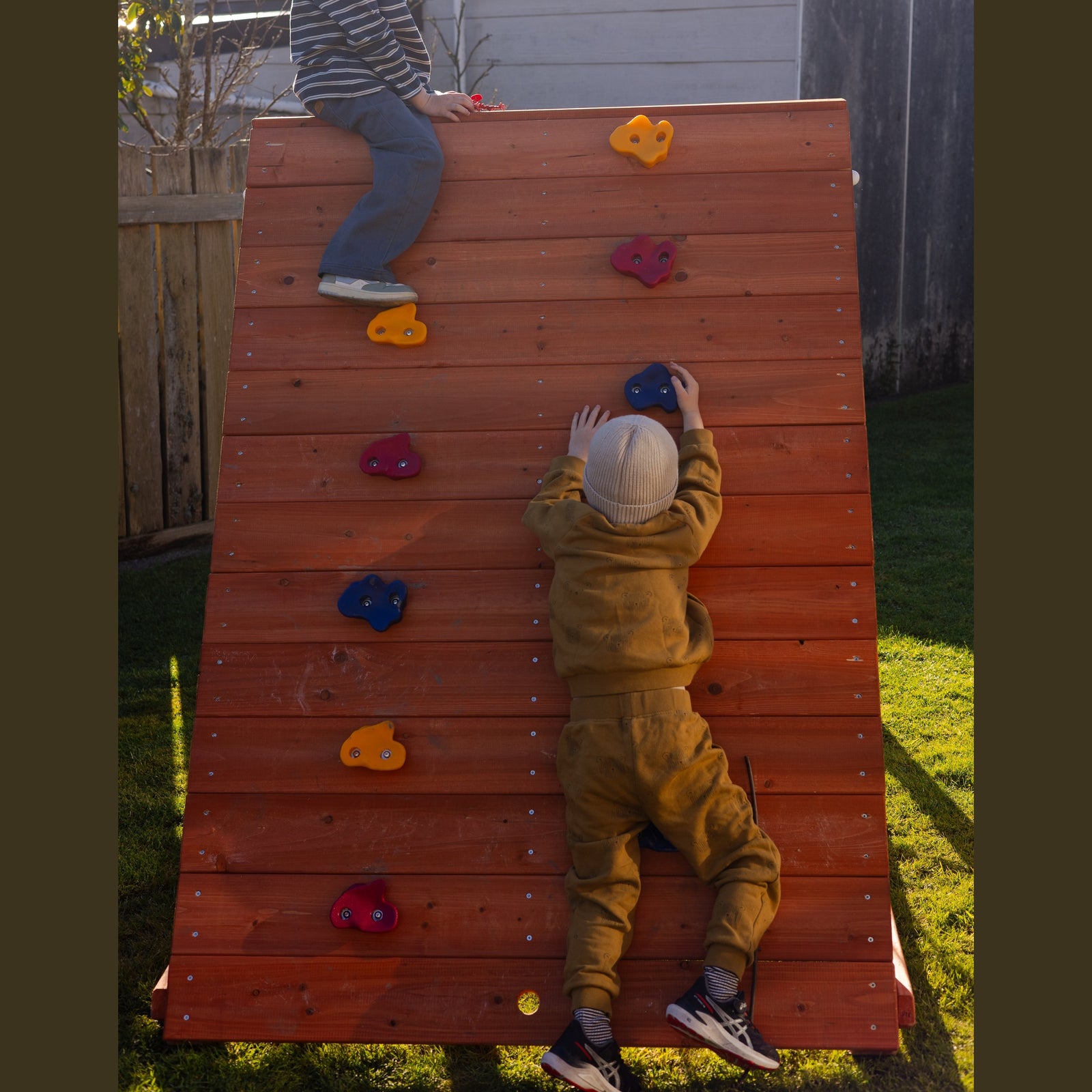 Two children playing on a wooden climbing wall outdoors in a backyard. One child is sitting at the top of the wall, and the other is climbing up the wall using colorful hand and foot holds.