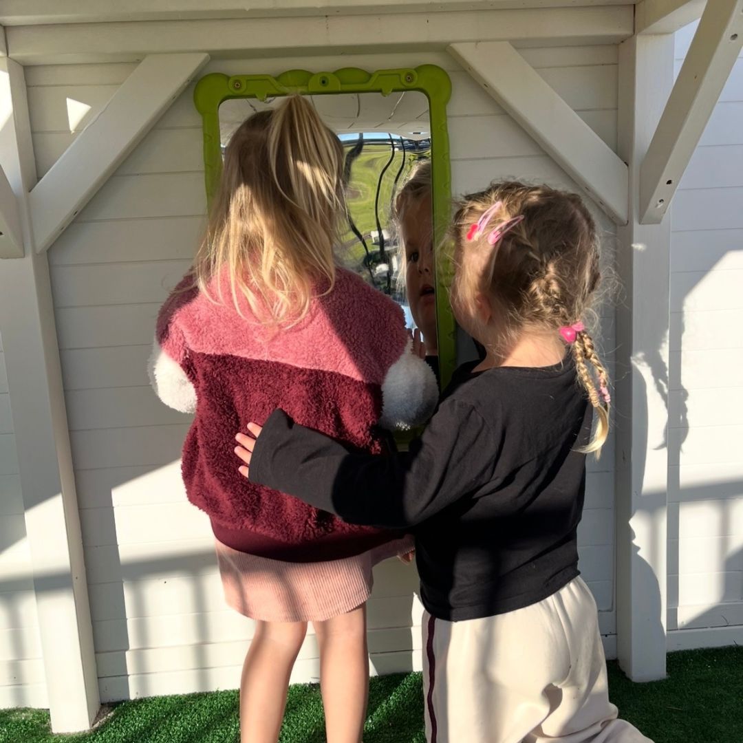 Two young girls standing in front of a white wooden playhouse wall, looking into a green-framed  funny faces mirror. One girl wears a pink and burgundy fuzzy top with a pink skirt, and the other wears a black long-sleeve top and cream skirt.