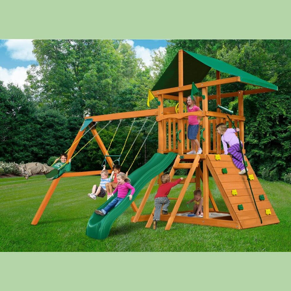 Children playing on a wooden playground set with a green slide and swings, surrounded by trees.
