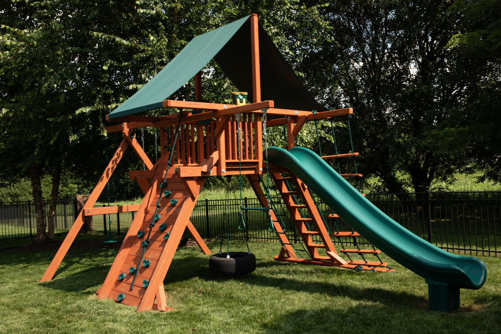 Children's wooden backyard playset with swings, a green slide, and climbing wall in a backyard setting.