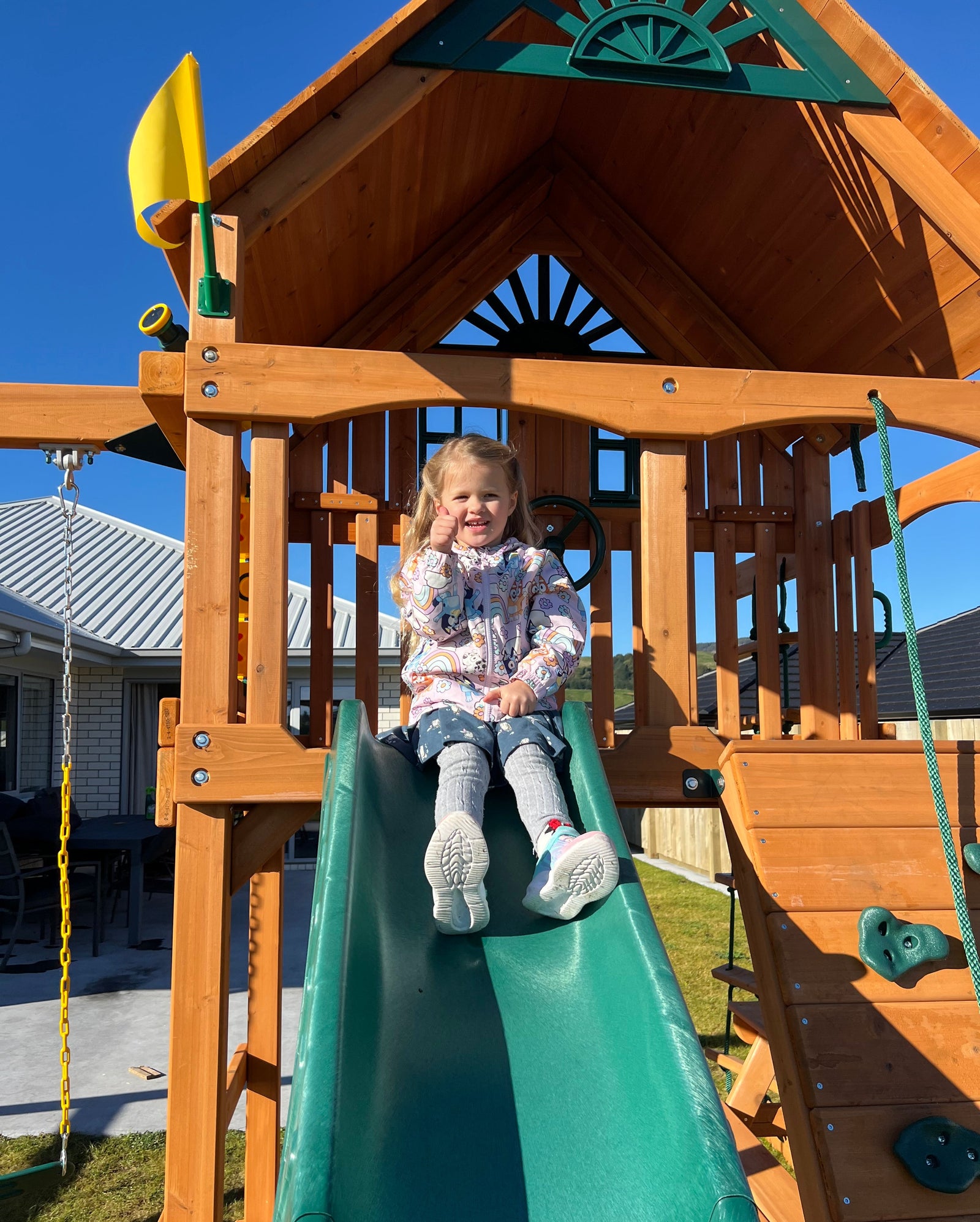 A young child slides down a green plastic slide on a large wooden playset in a backyard under a clear blue sky.