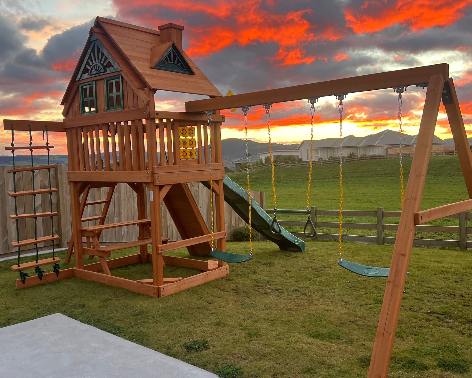 A wooden backyard playset featuring a two-story playhouse with a slide and ladder, connected to a swing set on a A-frame stand, set on a grassy lawn at sunset.