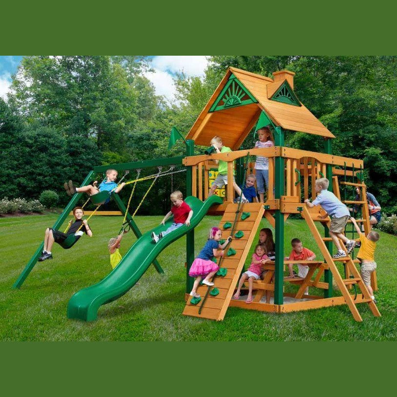 Children playing on a wooden playground set with green slides and swings in a backyard.
