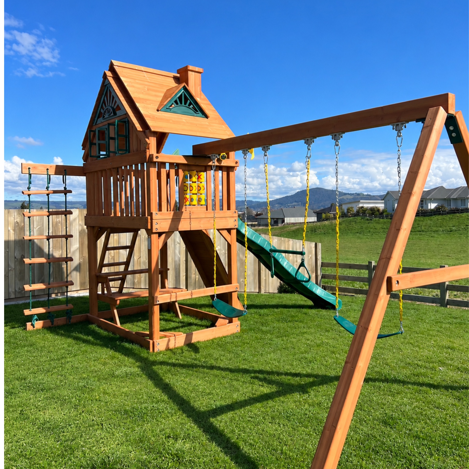 A large wooden backyard playset with a house, ladder, climbing wall, slide, and swing set, shown on a green lawn with a fence, hills, and blue sky in the background.
